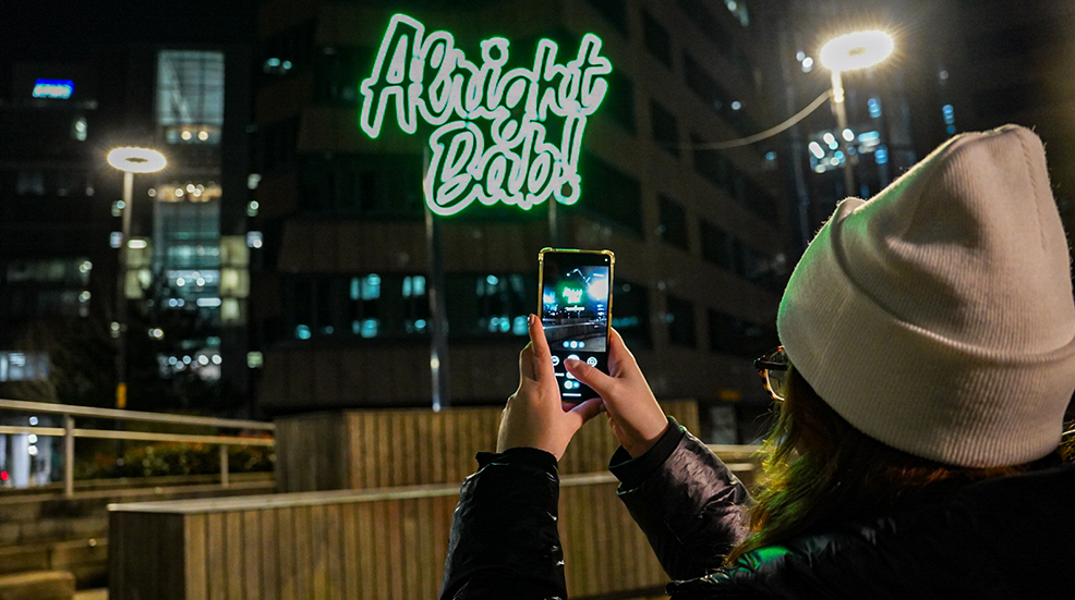 Woman taking photo with her smartphone at Birmingham Lights Festival neon sign reading 'Alright Bab!'